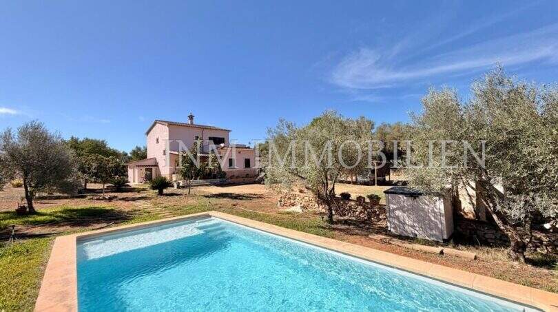 Außenansicht mediterrane Finca Algaida mit Pool, Olivenbäumen und rosa Haupthaus unter blauem Himmel.