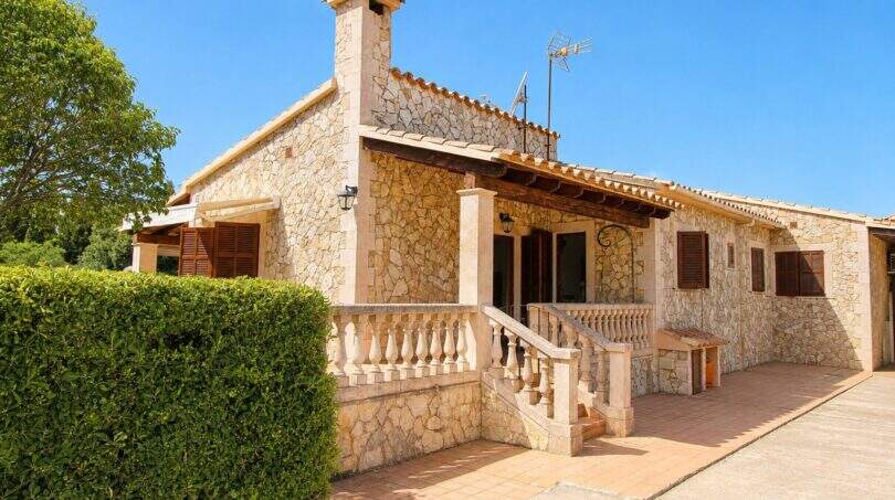 Außenansicht der Mallorca-Finca mit Natursteinmauern, Terrassenaufgang und Holzveranda unter blauem Himmel.