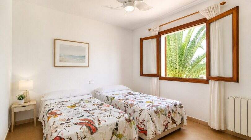 Helles Schlafzimmer der Finca Mallorca mit zwei Einzelbetten, Blumendecken, Deckenventilator und Palmenblick durchs Holzfenster.