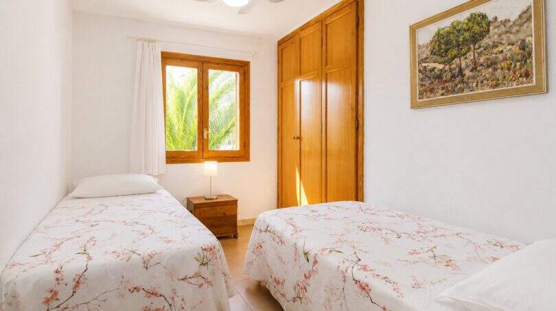 Helles Schlafzimmer der Finca mit zwei Einzelbetten, Blumendecken, Einbauschrank und Fenster mit Palmenblick.