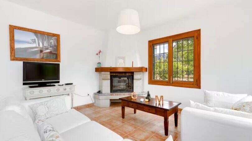 Helles Wohnzimmer der Finca mit weißen Sofas, Kamin, Holztisch und mediterranem Fensterblick ins Grüne.