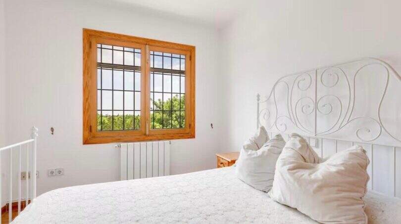 Helles Schlafzimmer der Finca mit weißem Schmiedeeisenbett, Holzfenster und Gartenblick in Palma de Mallorca.