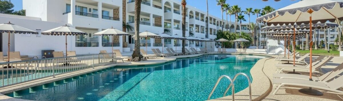 Piscina del hotel con tumbonas y sombrillas, fachada blanca con balcones y palmeras en Portocolom