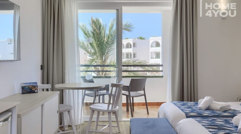 Habitación hotelera luminosa con balcón, vista a las palmeras, mesa de comedor y acentos azules en Portocolom