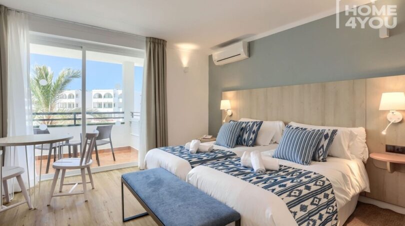 Habitación hotelera luminosa con camas dobles, decoración azul y blanco, aire acondicionado y balcón con vistas a palmeras en Portocolom