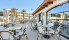 Terraza hotelera soleada con elegantes sillas de bistró, sombrillas y vista del área de piscina y edificio hotelero en Portocolom.