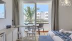 Habitación hotelera luminosa con balcón, vistas a palmeras, mesa de comedor y ropa de cama azul y blanca en Portocolom