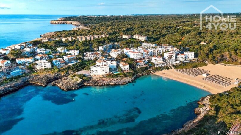 Vista aérea del apartahotel Portocolom: edificios blancos, bahía turquesa, playa de arena con sombrillas, vegetación exuberante.
