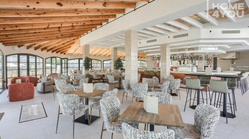 Restaurante hotelero luminoso con techo de vigas de madera, muebles modernos, bar y grandes ventanales en Portocolom