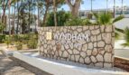 Señal de entrada del Hotel Wyndham Portocolom con muro de piedra natural, palmeras y edificio hotelero blanco de fondo