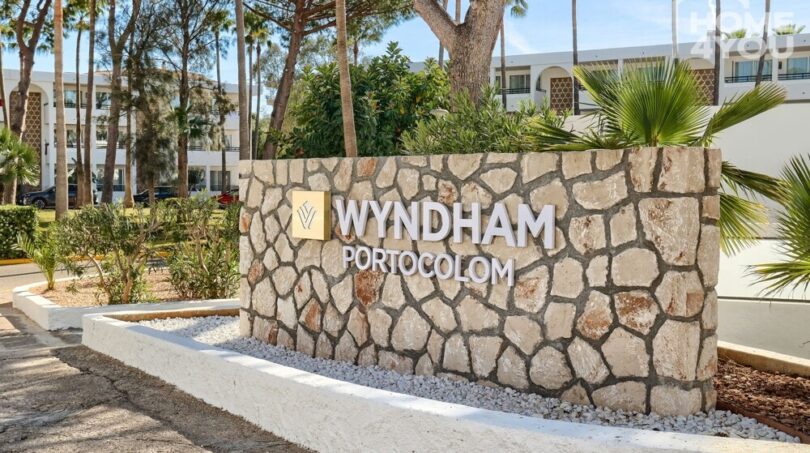 Señal de entrada del Hotel Wyndham Portocolom con muro de piedra natural, palmeras y edificio hotelero blanco de fondo