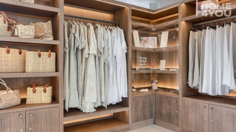 Elegante vestidor con estantería de madera, armarios empotrados iluminados, ropa blanca y bolsos de mimbre en el apartahotel Portocolom