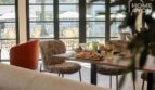 Interior de restaurante moderno con terraza, sillas elegantes, mesa servida con comidas, área de piscina al fondo.