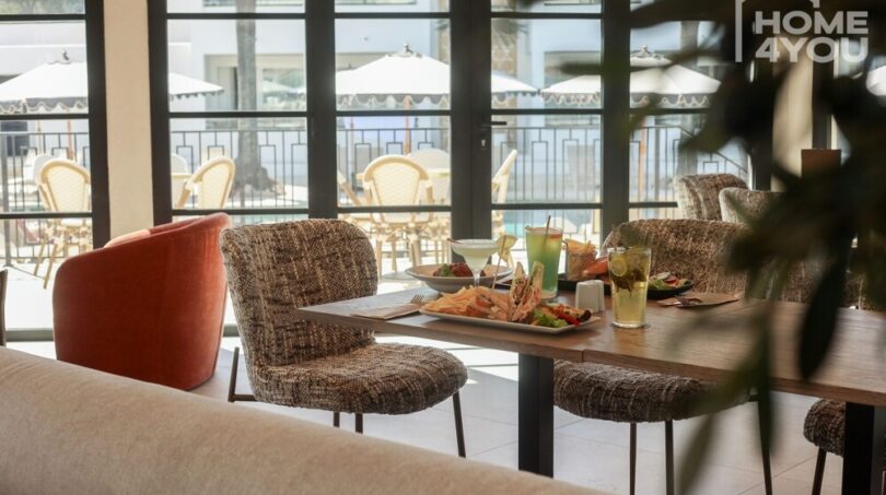 Interior de restaurante moderno con terraza, sillas elegantes, mesa servida con comidas, área de piscina al fondo.