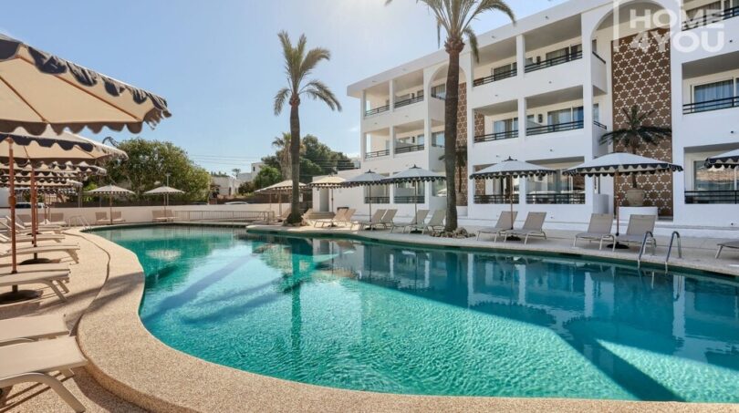 Complejo hotelero en Portocolom con piscina curva, tumbonas, sombrillas y fachada blanca con balcones