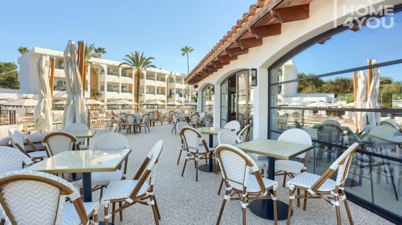 Terraza hotelera soleada con sillas de bistró, sombrillas, vista a la piscina y arquitectura mediterránea en Portocolom