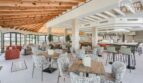 Restaurante hotelero luminoso con techo de vigas de madera, sillones modernos, bar y grandes ventanales en Portocolom