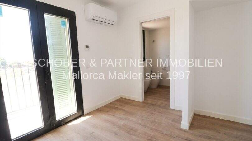 Dormitorio claro con suelo de madera, aire acondicionado, acceso al balcón y baño adyacente en apartamento esquinero de nueva construcción Cala Millor