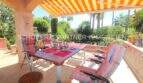 Terraza cubierta con mesa de comedor, tumbonas y vela solar naranja, rodeada de exuberante vegetación en Cala Millor.