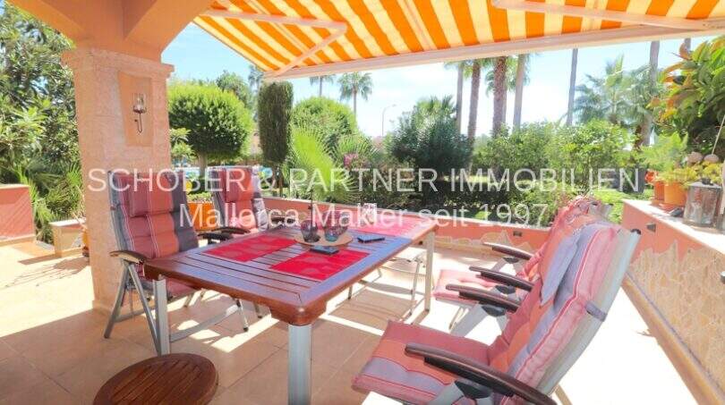 Terraza cubierta con mesa de comedor, tumbonas y vela solar naranja, rodeada de exuberante vegetación en Cala Millor.