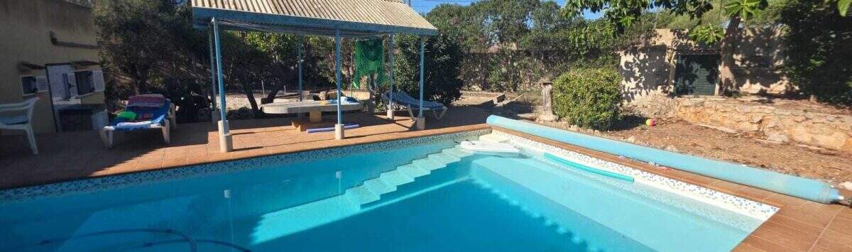 Outdoor pool of the finca in Cala Millor with sun terrace, pergola and lush greenery under blue sky.