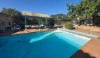 Outdoor pool of the finca in Cala Millor with sun terrace, pergola and lush greenery under blue sky.