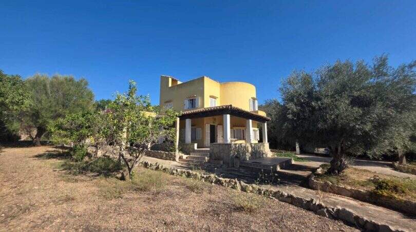 Exterior view of the yellow finca in Cala Millor with round tower, olive trees and large property under blue sky.