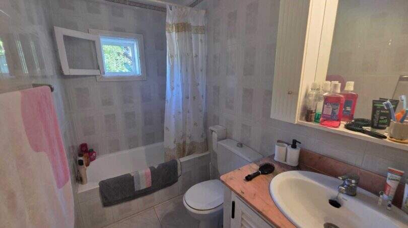Bright bathroom of the finca in Cala Millor with bathtub, shower curtain, toilet and washbasin with wooden vanity.