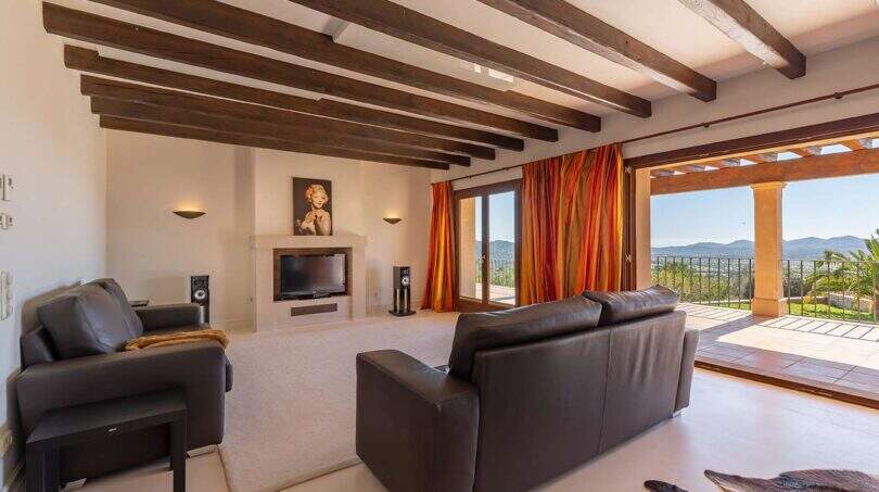 Bright living room of Felanitx finca with wooden beam ceiling, leather sofas, fireplace and panoramic view of the mountains.