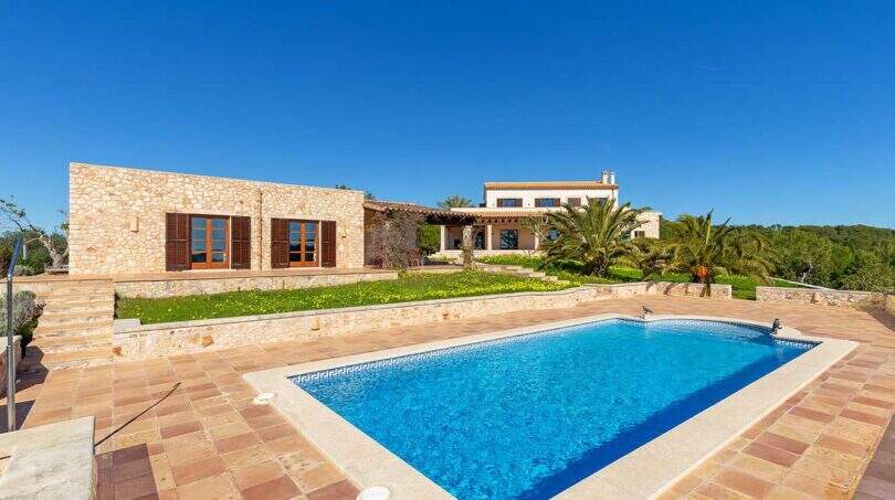 Exclusive finca in Felanitx with large swimming pool, natural stone walls, palms and well-maintained garden under blue sky.