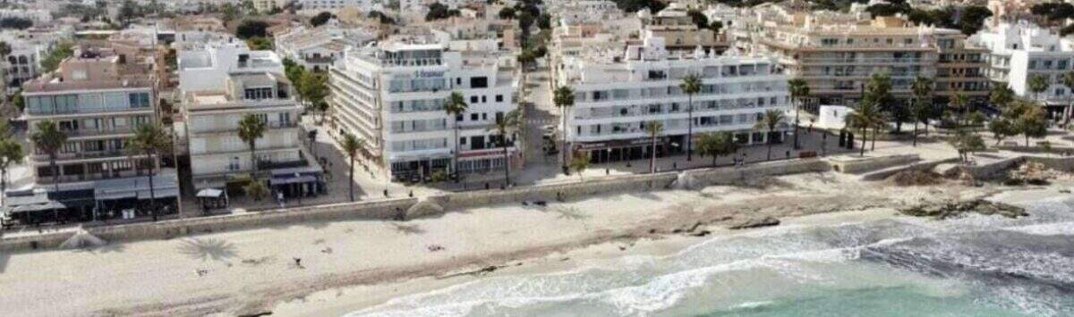 Luftaufnahme Strandpromenade Cala Millor mit weißen Gebäuden in erster Meereslinie, türkisem Wasser und Bergen im Hintergrund.