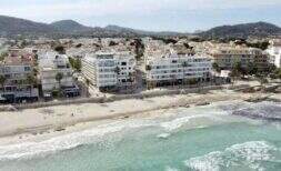 Luftaufnahme Strandpromenade Cala Millor mit weißen Gebäuden in erster Meereslinie, türkisem Wasser und Bergen im Hintergrund.