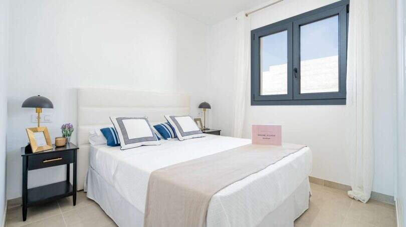 Dormitorio luminoso de la villa nueva en Cala Romántica con cama doble, mesitas de noche oscuras y ventana gris.