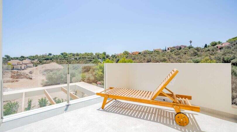 Azotea soleada de la villa nueva en Cala Romántica con tumbona de madera, barandilla de cristal y vistas a las colinas.