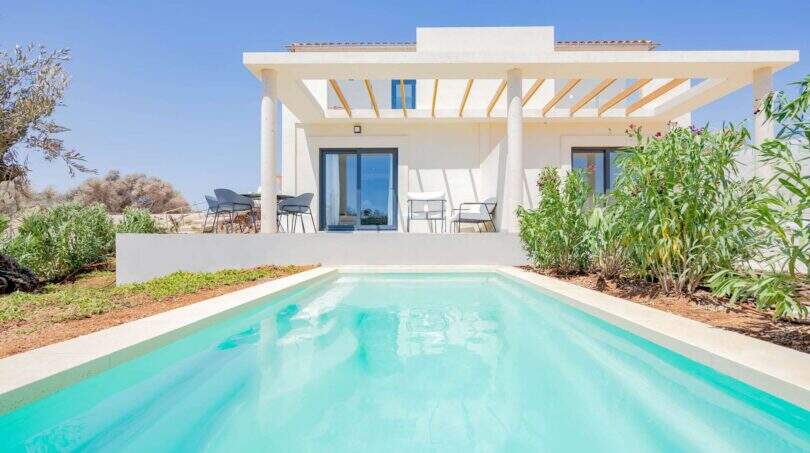 Vista exterior de la villa nueva en Cala Romántica con piscina privada, pérgola y jardín mediterráneo.