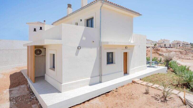 Vista exterior villa nueva Cala Romántica: casa de dos pisos blanca con techo de terracota, puerta de madera y terraza pavimentada.