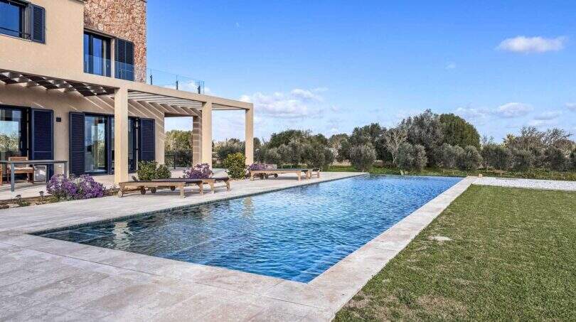 Luxusfinca Ses Salines: Rechteckiger Außenpool mit Pergola, Sonnenliegen und Olivenbäumen unter blauem Himmel.