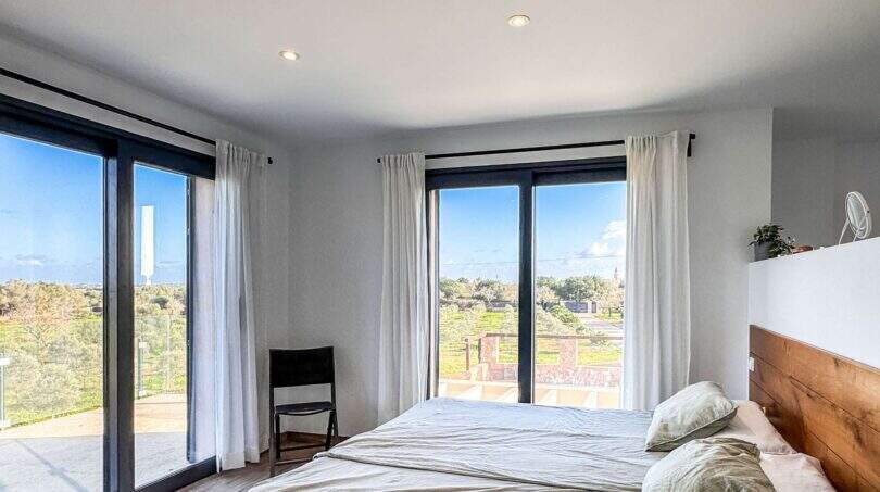 Helles Schlafzimmer der Luxusfinca Ses Salines mit Doppelbett, Holzkopfteil und Panoramafenstern mit Gartenblick.