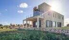 Außenansicht der Luxusfinca Ses Salines: Steinhaus mit Glasbalkon, überdachter Terrasse und grünem Garten bei Sonnenschein.