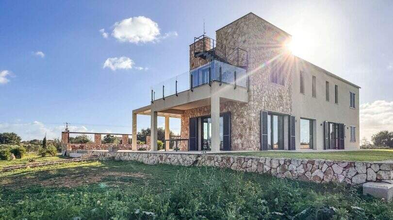 Außenansicht der Luxusfinca Ses Salines: Steinhaus mit Glasbalkon, überdachter Terrasse und grünem Garten bei Sonnenschein.