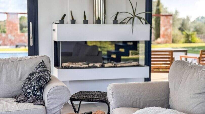 Modernes Wohnzimmer der Luxusfinca mit elegantem Glaskamin, Loungessofas und Blick in den mediterranen Garten.