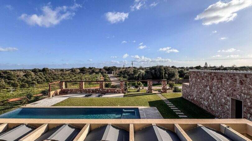 Luxusfinca Ses Salines: Pool mit Meerblick, Natursteinmauer und weitläufigem Garten unter blauem Himmel.