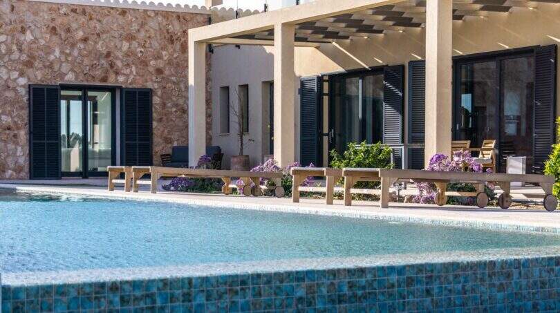 Luxusfinca Ses Salines: Pool mit Natursteinmauer, Holzliegen und überdachter Terrasse mit Blumenbeeten.