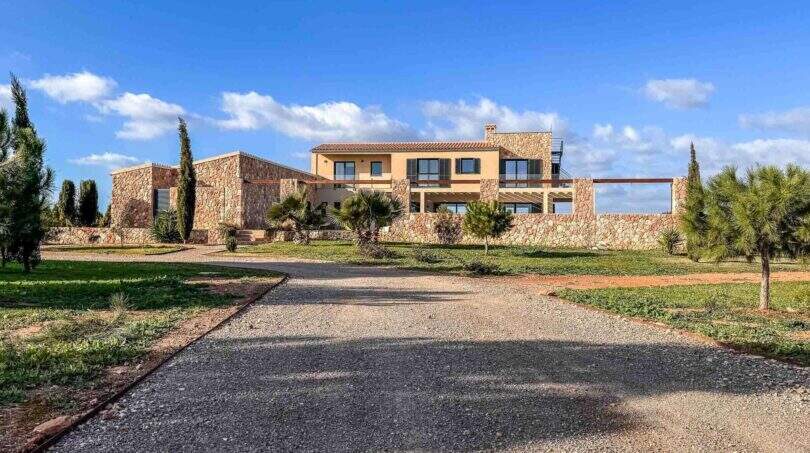 Außenansicht mediterrane Luxusfinca Ses Salines mit Natursteinmauern, Auffahrt und Gartenanlage unter blauem Himmel.