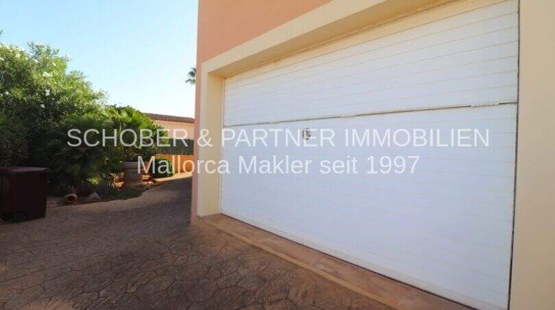 Garage mit weißem Rolltor am Einfamilienhaus in Sa Coma, gepflasterter Zufahrtsbereich mit Gartenblick.