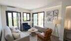 Área de estar clara del apartamento esquinero de nueva construcción en Cala Millor con sofá moderno, sillón de ratán y puertas balconeras.