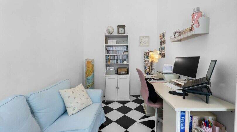 Despacho para trabajar en casa con piso de tablero de ajedrez, sofá azul, estantería blanca y escritorio con dos monitores en casa urbana Felanitx
