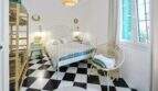 Dormitorio luminoso con piso de tablero de ajedrez, cama de hierro blanco, silla de ratán y persianas de color turquesa en casa urbana Felanitx