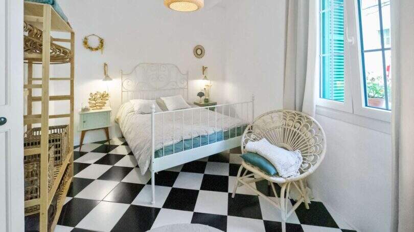 Dormitorio luminoso con piso de tablero de ajedrez, cama de hierro blanco, silla de ratán y persianas de color turquesa en casa urbana Felanitx
