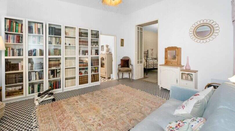Sala de estar luminosa con gran pared de libros, piso de baldosas con patrón, alfombra vintage y sofá azul claro en casa urbana Felanitx